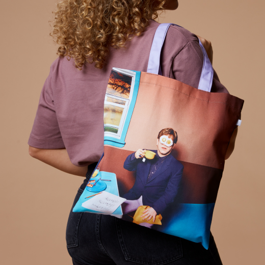 A woman wears a tote back featuring an photograph of a man sitting, holding a cup of coffee.